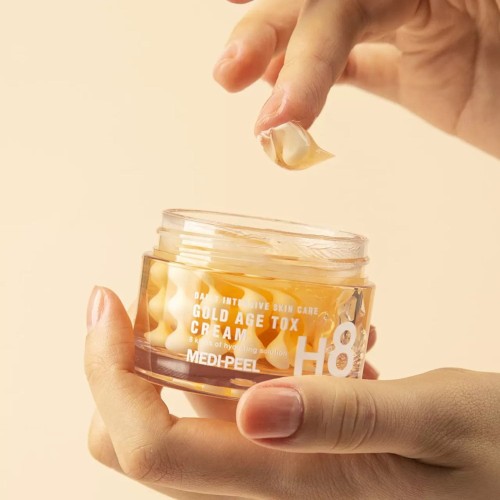 MEDIPEEL Gold Age Tox Cream krem przeciwstarzeniowy w kapsułkach - zbliżenie na żelowe kapsułki trzymane w dłoni nad otwartym słoikiem z kremem, na beżowym tle.